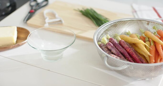 Camera is panning, revealing colander of multicolored carrots, glass bowl, butter, chives for prep