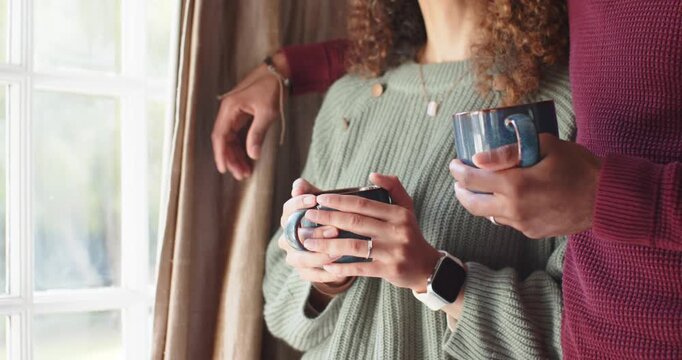 Diverse couple by window in knits cupping mugs, after woman glancing up, man speaking, connecting