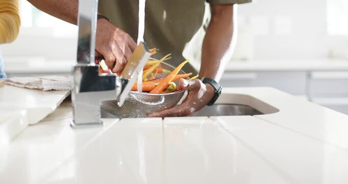 Man is rinsing carrots, roots at sink in colander as chrome faucet runs, removing dirt