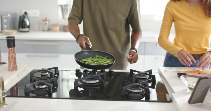 Couple cooking at kitchen island, lit flame urging man flipping pan of green beans, woman prepping