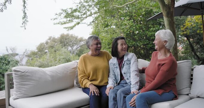 Diverse female friends sitting on patio sectional, two joining, laughing, catching up