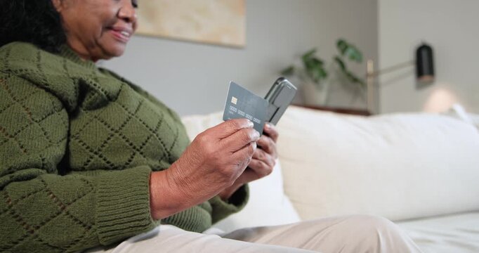 Senior African American woman wearing green sweater on sofa checking card, tapping phone to pay