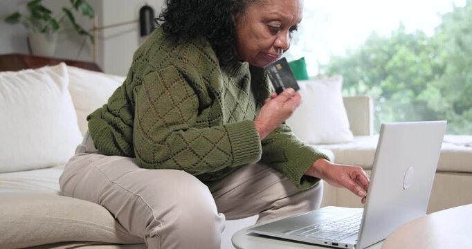 African American senior woman leaning at home on sofa, typing on laptop, entering card for payment