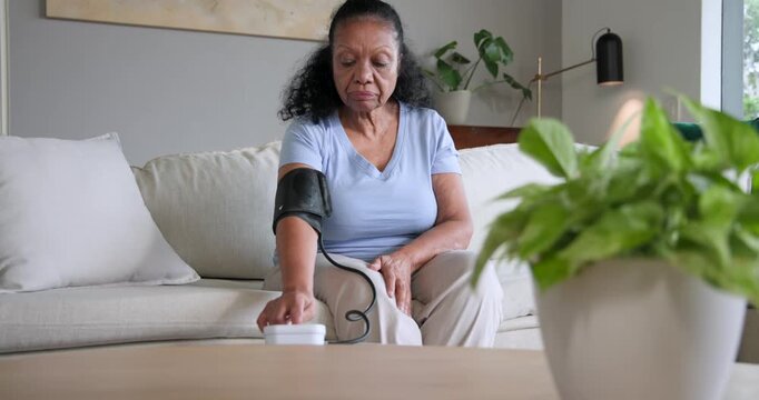 Senior African American woman preparing for BP check, placing cuff and using white monitor on sofa