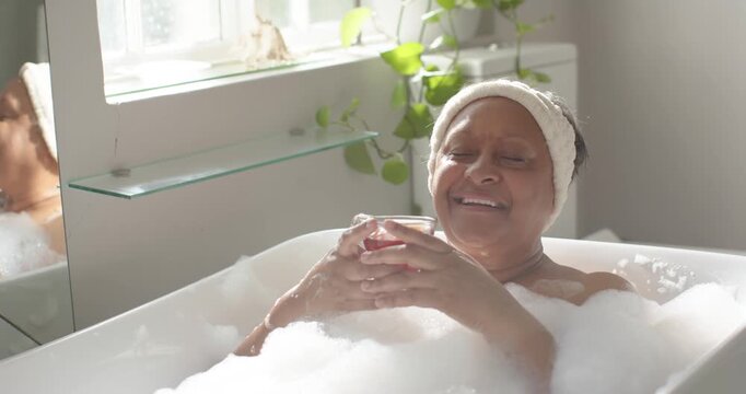 Mature African American woman settling into bathtub foam, holding red drink and resting by window