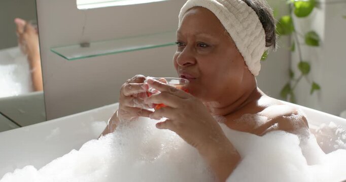Mature African American woman settling into bathtub foam wearing headband, sipping drink for calm