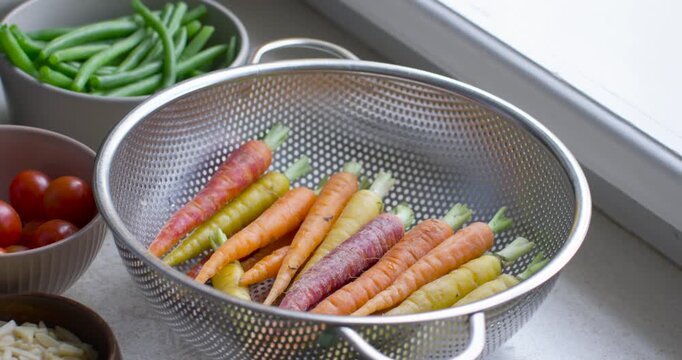 Camera reframing and zooming is centering metal colander and is showing heirloom carrots at window