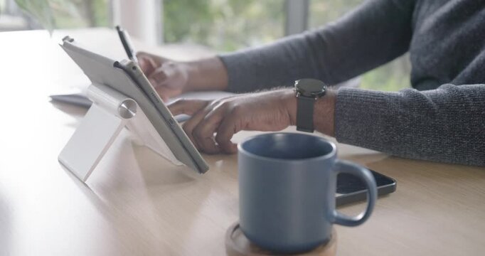 African American man tapping tablet to reference content and writing in notebook at home workspace