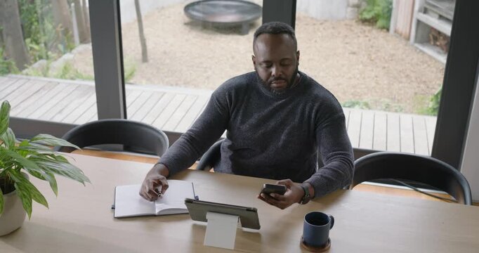 African American man checking phone writing in notebook and tapping tablet during work call at home