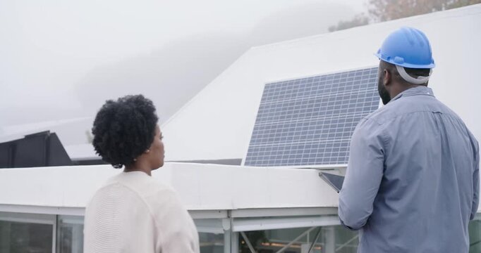 African technician with hard hat, gloves showing tablet and client checking solar panels on rooftop