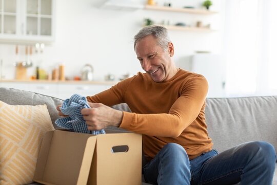 Happy Elderly Caucasian Man Smiling While Unboxing Clothes From A Package At Home, Sitting Comfortably On Couch In Living Room, Experiencing Joy And Excitement. Retail Experience Concept.