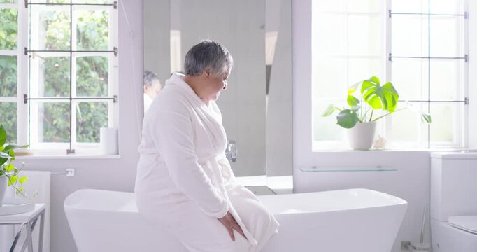 Senior African American woman sitting tub rim in bathroom bracing, fixing white robe, looking right
