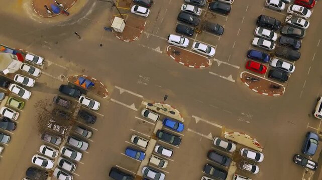 Aerial view of a busy parking lot with organized rows of parked vehicles and ongoing urban traffic in daylight.