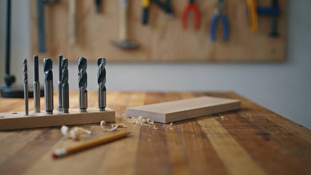 Drill bits stand in an organized row on a wooden workbench with a pencil and wood shavings in the background.
