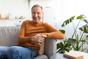 Fototapeta premium Smiling Elderly Caucasian Man Sitting Comfortably On Couch With Hand On Knee In Cozy Living Room. Cheerful Senior Enjoying Relaxed Moment At Home, Highlighting Comfortable Lifestyle Concept.