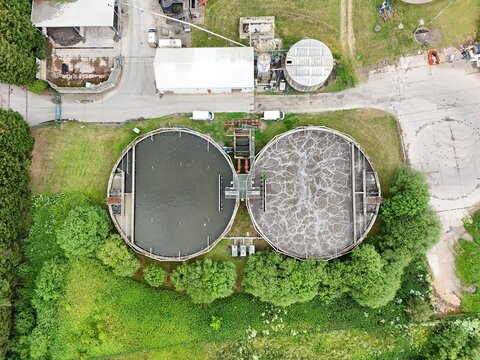Aerial view of a wastewater treatment plant with two large circular sedimentation tanks and industrial infrastructure surrounded by green vegetation in the United Kingdom.