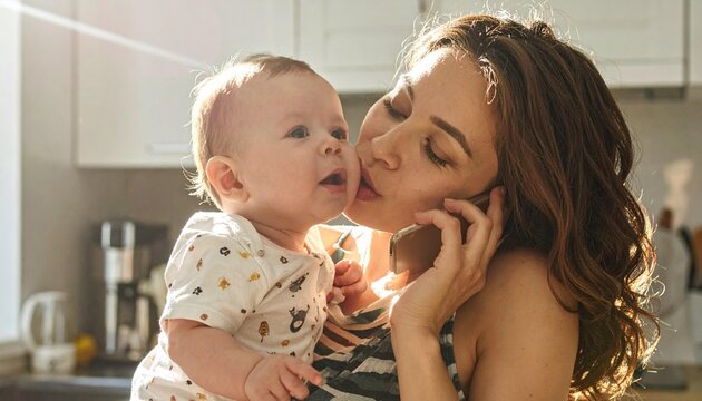 Mother on phone kiss baby gently in sunlit kitchen