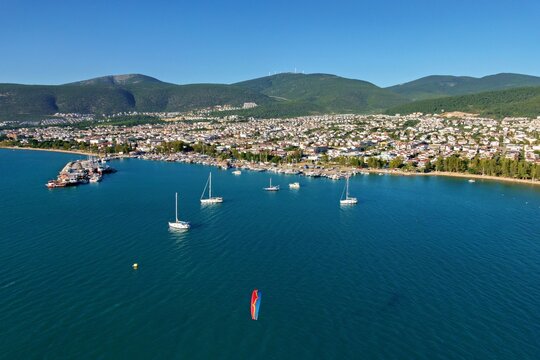 Aerial view of the coastal town of Akbuk with sailboats moored in the bay, a paraglider flying over the blue sea, and hills in the background in Akbuk, Aydin, Turkey.