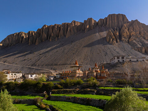 Aerial view of a traditional Buddhist monastery nestled at the base of dramatic, rugged mountain cliffs, surrounded by vibrant green terraced fields under a clear blue sky in Nepal.