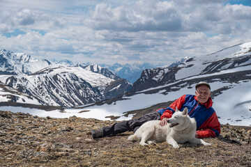 Front view of happy senior man with dog resting on mountain hill. Active elderly traveler with husky enjoying trekking travel adventure in remote snowy wilderness landscape. © ANDREY