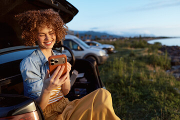 Naklejka premium Young woman smiling outdoors by the car trunk enjoying a casual lifestyle wearing denim and sunlight captured in a candid moment during a relaxed coastal outing