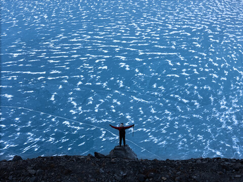 Aerial view of a person in a red jacket standing on a rocky outcrop overlooking a vast frozen blue lake with intricate ice textures in Nepal.