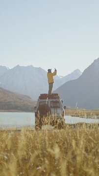 Traveler in a yellow jacket standing on the roof of an off road SUV and taking a smartphone photo by a mountain lake in a remote alpine valley. Golden grass, snowy peaks, and bright sunlight create a