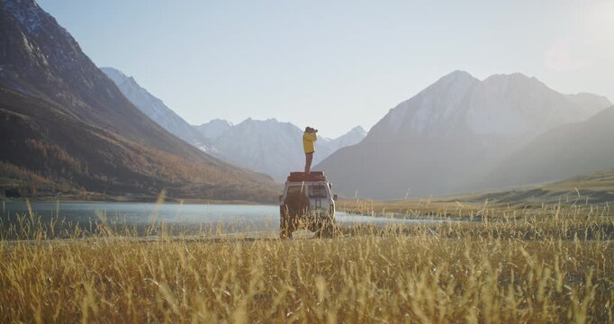 Traveler with a camera standing on the roof of an off road vehicle near a mountain lake surrounded by snowy peaks and a wide alpine valley. Warm sunlight, golden grass, and open wilderness create a