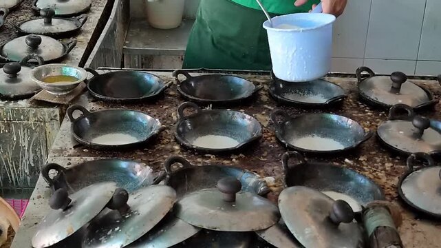 Close-up of a vendor pouring rice flour and coconut milk batter into multiple small hot woks with a ladle while preparing traditional Indonesian Surabi Solo cakes.