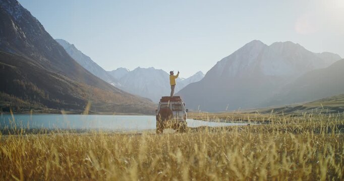 Man in a yellow jacket standing on the roof of an off road vehicle and shooting a phone picture by a mountain lake with snowy peaks in the background. Wide natural landscape with dry grass and bright