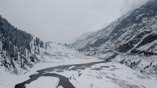 Aerial view of snow-covered mountains and a winding river flowing through a winter landscape under an overcast sky in Babusar, KPK, Pakistan.