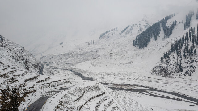 Aerial view of the snow-covered Babusar Pass landscape with a winding mountain road, a frozen riverbed, and pine trees under a cloudy sky in Babusar, KPK, Pakistan.