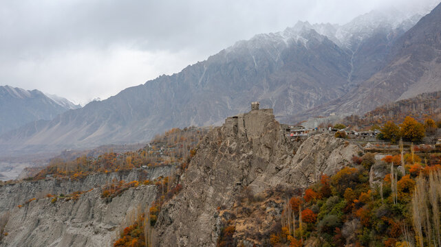 Aerial view of Altit Fort perched on a rocky cliff surrounded by autumn trees and snow-capped mountains in Altit, Gilgit, Pakistan.
