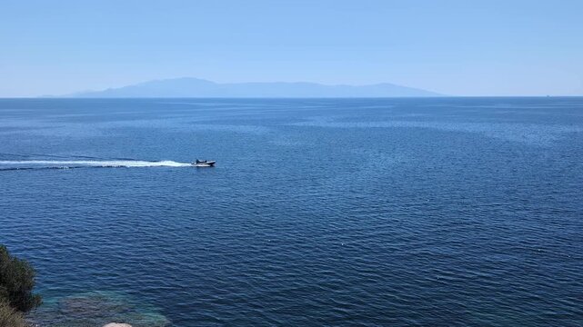 A speedboat speeds across a calm, blue sea under a clear sky, with distant mountains in the background. The water reflects the sunlight, creating a shimmering effect.