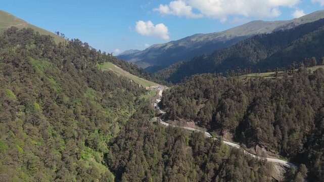 Aerial view of dirt road winding through dense green forest in mountain valley, remote infrastructure b-roll