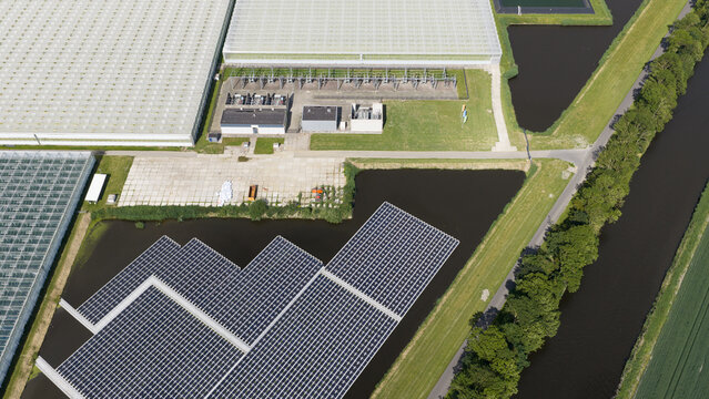 Aerial view of floating solar panels installed on a pond adjacent to large industrial greenhouses in Middenmeer, North Holland, Netherlands.