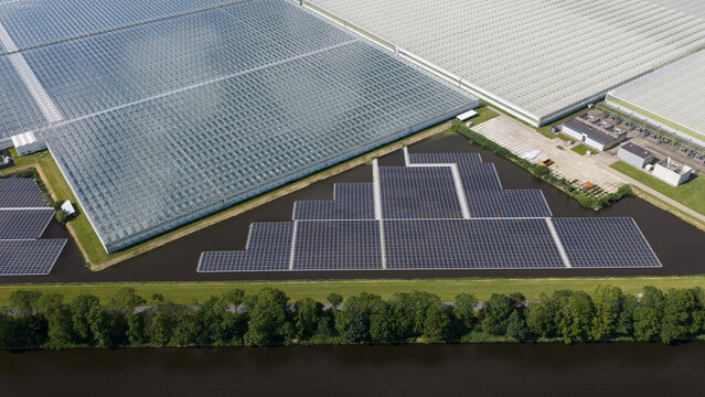 Aerial view of a floating solar farm on a pond adjacent to large glass greenhouses in Middenmeer, North Holland, Netherlands.