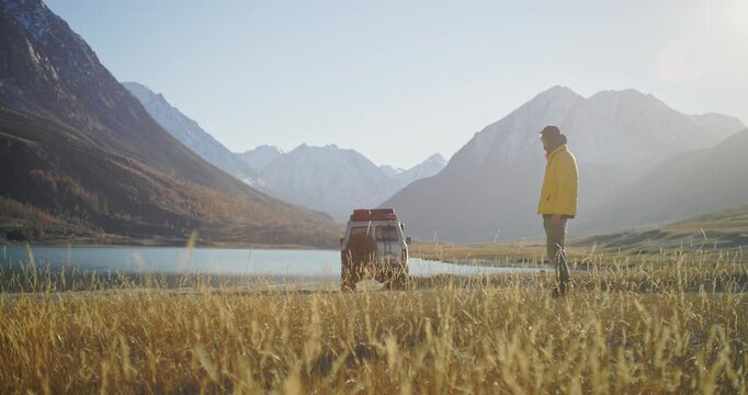 Traveler with a camera standing near an off road car by a calm mountain lake surrounded by snowy peaks and a wide alpine valley. Warm sunlight, golden grass, and open wilderness create a strong