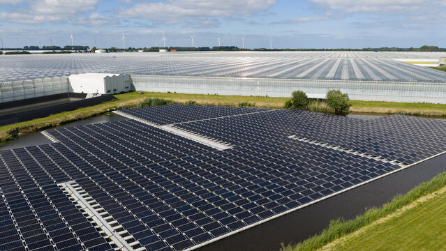 Aerial view of floating solar panels on a canal adjacent to a vast greenhouse complex under a blue sky with wind turbines in the distance Middenmeer, North Holland, Netherlands.