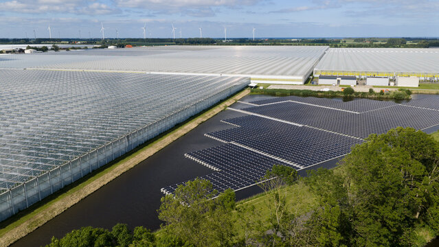 Aerial view of a large greenhouse complex adjacent to a floating solar panel array on a canal in Middenmeer, North Holland, Netherlands.