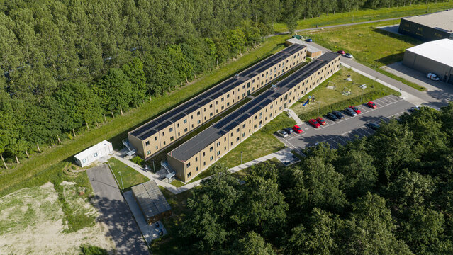 Aerial view of a long, modern building complex with solar panels on the roof, adjacent to a parking lot and surrounded by trees in Middenmeer, North Holland, Netherlands.