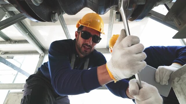Two industrial technicians wearing helmets and safety gear tightening a pipeline connection with a large wrench inside mechanical facility infrastructure.