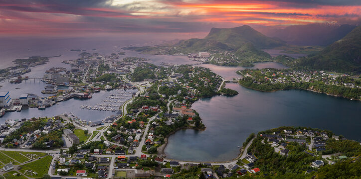 Aerial view of Svolvaer, Lofoten Islands, Norway, at sunset. Houses cluster by marinas, boats line docks, curving roads link islets, and a dark mountain rises behind.