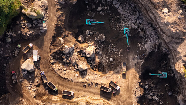 Aerial view of excavators and dump trucks working in a stone quarry with dirt roads and large boulders in Indonesia.