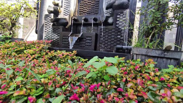 A video shot of a decorative garden featuring a dark stone water fountain with multiple spouts flowing into a basin.