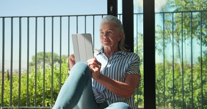 Senior woman sitting at sliding glass door swiping tablet, smiling as sunlight drawing gaze outside
