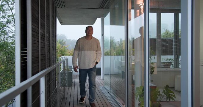 Senior Asian man emerging indoors walking onto deck keeping hands in pockets viewing misty hills