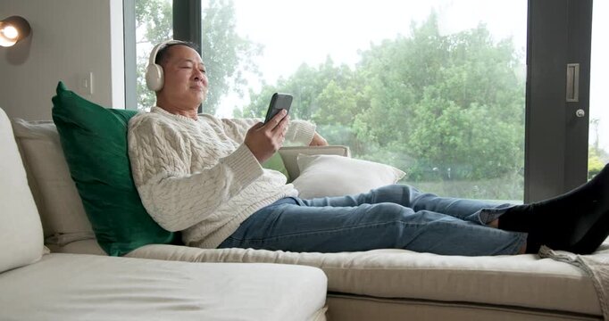 Senior Chinese man scrolling phone on sofa by window, removing headphones then standing, gazing out