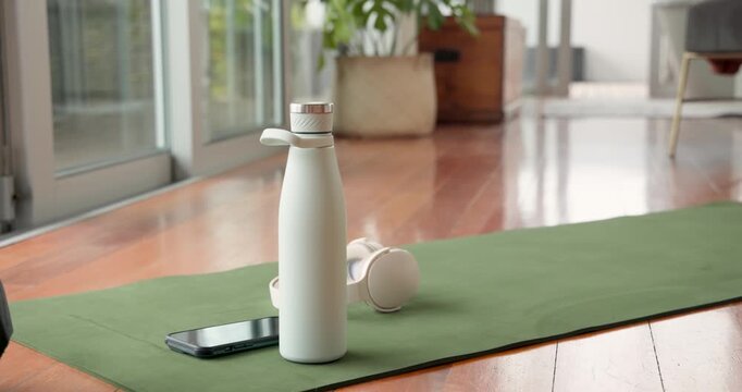 Camera is shifting right and down, white bottle is standing on green mat showing headphones, phone