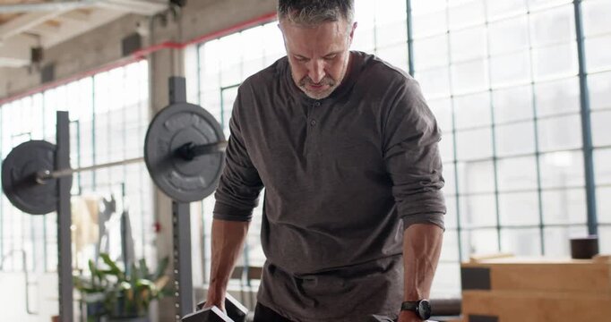 Middle-aged man holding hex dumbbells, curling, lowering to bench at gym, preparing for next set